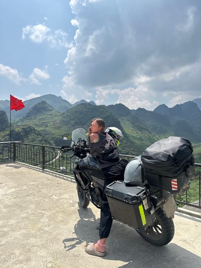 A person is sitting on a parked motorcycle on a concrete platform, with mountains and a partly cloudy sky in the background. The motorcycle is black with panniers, a top case, and a helmet strapped to the back. The person is wearing riding gear, including a jacket and a helmet. A red flag is visible in the lower left corner of the image. The location is Lũng Phìn Commune, Vietnam.