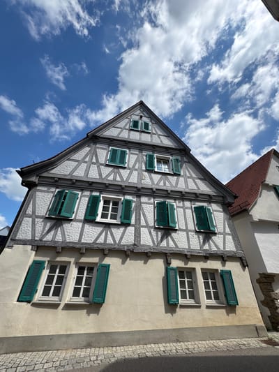 This is a low-angle, vertical outdoor photograph of a traditional half-timbered building in Bietigheim-Bissingen, Germany. The building has a white plastered facade with dark wooden beams forming diagonal and horizontal patterns. There are multiple windows, most of which are covered by dark green shutters that are partially open. The roof is gabled and tiled. The sky above is blue with scattered white clouds. To the right of the half-timbered building, a portion of another building with a red tiled roof and white facade is visible. The foreground shows a cobblestone street. The image captures a historic architectural style under a partly cloudy sky.