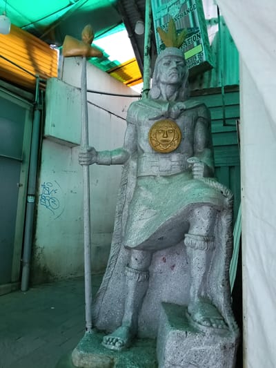 The image shows a silver-colored statue of a person with indigenous features and garb. The statue holds a spear-like staff in its left hand and has a large, circular, golden sun-like emblem on its chest. It is wearing a crown and stands on a rough-hewn stone pedestal. The setting is an urban alleyway, lined with walls of concrete and corrugated metal, and covered with green and yellow translucent roofing. The backdrop includes a sign for "Money Exchange." Graffiti is visible on the left wall. This is likely in or near the city of Machu Picchu, Peru, given the statue's indigenous imagery.