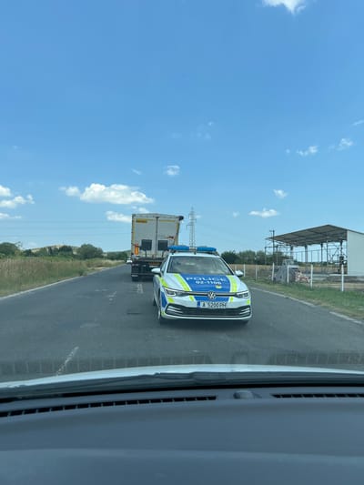 The image depicts a police car and a truck on a paved road with a grassy verge. The police car is a white Volkswagen Golf with blue and yellow markings and the word "POLICE" visible on its side. Its license plate reads "A 5200 PH". The truck is white and has "WILLTON" written on its side. In the background, there's a large, open-sided structure with a metal frame and roof, possibly a warehouse or agricultural building. Further back, there are trees and a hill. Power lines and a pylon are also visible. The sky is bright blue with scattered white clouds, suggesting it is daytime with good weather. The scene appears to be on the outskirts of Kableshkovo, Bulgaria, given the context. The image seems to be taken from inside another vehicle, possibly looking out through a windshield or window. No people are clearly visible.