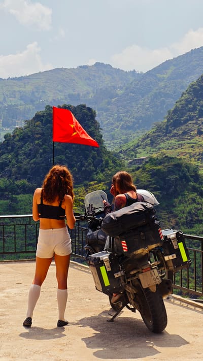 Two women stand near a parked touring motorcycle on a concrete platform with a black metal railing. The motorcycle is heavily laden with luggage and panniers. A red flag with a yellow star flies from a pole nearby. The woman on the left has red curly hair, wears a black top, white shorts, and knee-high socks with patterned shoes. The woman on the right has brown hair, a tattoo on her arm, and is looking at a phone. The motorcycle's license plate is visible. Behind them is a mountainous landscape covered in dense green vegetation, under a sky with some clouds. The scene is likely in Lũng Phìn Commune, Vietnam.
