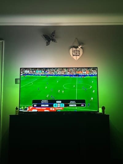In the city of Marten, Bulgaria, an indoor scene shows a large flat-screen television displaying a soccer match. The score displayed on the screen indicates England is playing Croatia, with England leading 4 to 2. The match appears to be nearing its end, with the time showing 86:53. The television is positioned on a dark cabinet, which is lit from behind by green ambient lighting. Above the television, on the wall, are two decorative items: a black butterfly with rhinestones and a wooden heart-shaped plaque with the words "with love" carved into it. The wall itself is painted a dark grey color and a white crown molding runs along the top of the wall.
