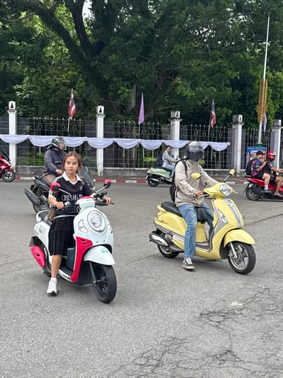 In this image, two people are seen riding scooters in Suthep, Thailand. The first scooter, a yellow model, is ridden by a person wearing a light-colored jacket and blue jeans, with a helmet on. A second person is partially visible on the back of this scooter. Behind them, another scooter, red in color, is also occupied by two individuals, one wearing a red helmet.
In the foreground, a white and red scooter is parked, and a young woman is seated on it, looking towards the camera. She is wearing a black and white athletic top and a black skirt, with white sneakers. A person wearing a helmet is seated behind her on the same scooter. The scooters are parked on a paved road, with a metal fence adorned with white draped fabric and small flags in the background. Lush green trees are visible behind the fence. The scene appears to be outdoors, possibly in a street or a public area, given the presence of the fence and road. The lighting suggests it might be daytime, with overcast conditions.
Visible text on the white scooter reads "CHULAKO." The overall mood seems casual, with the individuals appearing relaxed as they pose for the photo or are in transit. The location context is identified as Suthep, Thailand.