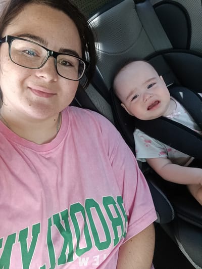 An overhead, medium shot shows a woman in a pink t-shirt with green lettering and a baby in a car seat. The woman is wearing glasses and smiling at the camera. The baby is crying and looking away from the camera. The car seat is black with gray accents. The scene appears to be inside a car.