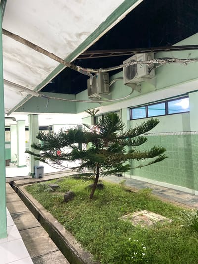 The media file depicts an outdoor scene, likely in Margoyoso, Indonesia, at an unspecified time of day that appears to be dusk or night, given the lighting conditions. The setting is an exterior corridor of a building, possibly a school or dormitory, with air conditioning units mounted on the wall. A narrow strip of green grass and a small tree are visible to the right, bordering a concrete pathway. The building's exterior is painted a light green and white, with visible pipes and wiring running along the walls. The overall mood is utilitarian and perhaps a bit neglected, with some of the pipes appearing worn or patched. There are no people visible in the image, and the focus is on the architecture and the surrounding vegetation.
