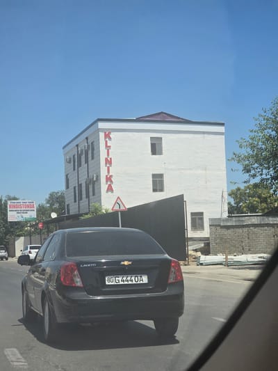 A black Chevrolet Lacetti sedan is driving on a paved road in Bukhara, Uzbekistan. The car has a license plate reading "80 G 444 OA UZ". In the background, a white, multi-story building with red lettering that spells "KLINIKA" is visible. There is also a billboard in the background with text in Uzbek, advertising medical tourism. The sky is clear and blue, and trees are present on the right side of the image. The scene appears to be a sunny day.