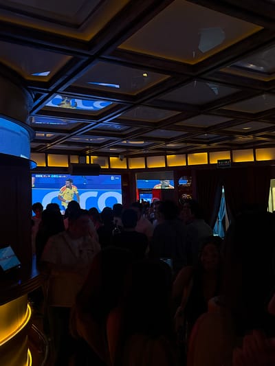 In an indoor establishment with a dark wood-paneled ceiling featuring recessed lighting and square panels, a crowd of people is gathered. The ceiling panels reflect light, and some show distorted images. A large screen at the front of the room displays a soccer match with a player in a yellow jersey. The bottom of the screen shows a game score. Some individuals in the crowd are looking at the screen, while others are engaged in conversation. The environment appears to be a bar or a sports viewing venue. The image is likely from San Pedro Garza García, Mexico, given the presence of "SALIDA" (Exit in Spanish) sign and the general ambiance.