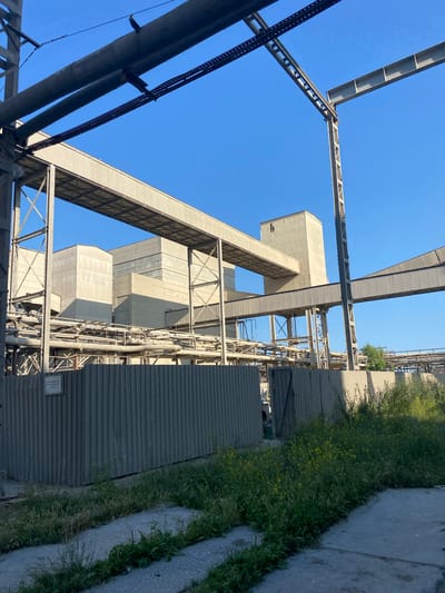 The image depicts an industrial facility with a clear blue sky overhead. The structures are characterized by large, concrete buildings, elevated conveyor belts, and extensive piping systems. A corrugated metal fence runs along the foreground, partially obscuring a section of the industrial complex. Overgrown vegetation, including tall grass and yellow wildflowers, grows around the base of the fence and on unpaved areas. The scene suggests a manufacturing or processing plant, possibly in a state of operation or disuse, situated in an outdoor environment.