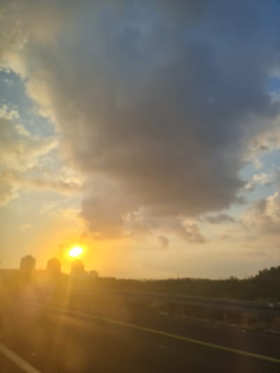 The image captures a blurred view of a sunset over a highway in Caesarea, Israel. The sun, a bright orb, is partially obscured by dark clouds and casts a warm, golden glow across the sky. Silhouetted buildings are visible on the horizon, suggesting an urban or suburban landscape. The highway itself is dimly lit, with a yellow line marking the lane. Vegetation lines the side of the road, and a hazy, possibly coastal, environment is implied. The overall impression is one of a dramatic and serene end to the day.