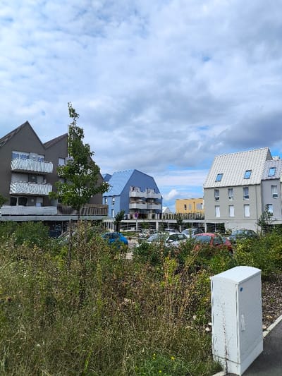 The image depicts a modern urban landscape in Wolfisheim, France, under a partly cloudy sky. In the foreground, a substantial area of unkempt, wild vegetation, primarily green and brown foliage, occupies the lower left and central portions of the frame. A white utility cabinet with a textured surface and a small blue circular emblem on its upper right side is positioned on a dark gray paved path in the lower right corner.
Behind the foreground vegetation, a paved parking lot is visible, containing multiple parked passenger vehicles of various colors, including a blue car, several white cars, and a red car. A brown, circular regulatory sign is present within the parking area.
The background features several multi-story buildings. On the left, a dark gray building with a distinct irregular peaked roofline and multiple balconies with white railings is prominent. A tall, slender tree with green leaves stands in front of this building. To the right of the dark gray structure, a bright blue building with a similar peaked roof and balconies is visible. Further to the right, a light gray building features a white, horizontally ribbed metal roof punctuated by multiple skylight windows. A yellow-colored building is partially visible in the far background between the blue and light gray structures. The sky overhead is largely covered by white and light gray clouds, with patches of lighter blue showing through. No people are present, and no actions or interactions are depicted.