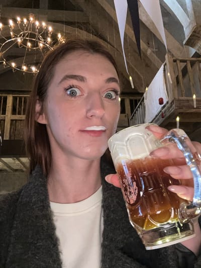 A female subject is depicted in a medium close-up, directly facing the camera with wide-open blue eyes and a line of white foam on her upper lip. She holds a clear, textured glass mug containing a dark beverage topped with a thick layer of white foam in her right hand, her nails manicured with a light polish. She wears a dark gray textured coat over a light-colored shirt. The background reveals an interior space with a high, vaulted wooden ceiling and exposed beams. Overhead, a large chandelier with multiple candle-like lights hangs, alongside several suspended individual floating candle lights and white and dark banners. A wooden balcony with a railing is visible in the background, from which a white fabric banner with red spherical accents hangs. The scene is located within Licheng District, China.