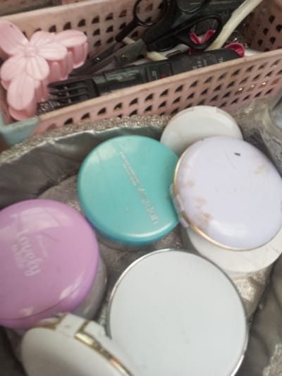 Several cosmetic compacts in pink, turquoise, and white with gold accents are scattered inside a silver-colored bag. The pink compact features "Lovely White" lettering. A pink plastic flower hair clip, scissors, and other small tools are placed in a pink plastic basket behind the compacts. The basket's design includes small, evenly spaced openings.