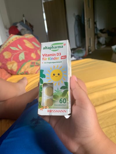 A person's hand is holding a box of "altapharma Vitamin D3 für Kinder" (for children) chewable tablets. The box is white with green and orange accents, featuring a cartoon sun and jungle leaves. Text on the box includes "Vitamin D3", "für Kinder", "mit Tropicalgeschmack" (with tropical flavor), "60 Kautabletten" (60 chewable tablets), and "ROSSMANN". Three yellow chewable tablets are visible through a clear window in the box. The background shows a blurred interior scene with yellow and red fabric. The image appears to be taken indoors during the day, likely in a home. The person's hand suggests they are about to take or have just received the vitamins.