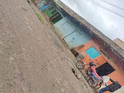 This image depicts an outdoor street scene in Juja ward, Kenya. The foreground and mid-ground consist of an unpaved, brownish-grey ground surface. To the right, a row of low-rise buildings is visible. The closest building is peach-orange, featuring a bright blue rectangular opening covered by a white metal security grid, and a darker, recessed opening. A partial inscription, "WANANITU," is visible on this building's facade. Behind it, additional structures with light blue, green, and red elements are discernible. Overhead, multiple parallel utility lines traverse an overcast, light grey sky. A red and blue motorcycle, equipped with a rear rack, is parked on the unpaved ground, oriented towards the right. A partially visible individual, dressed in blue trousers and a dark upper garment, stands adjacent to the motorcycle in the lower right portion of the frame. Their right arm is extended towards the motorcycle, holding or interacting with a white bag.
