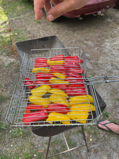In Vyshhorod, Ukraine, a metal grill basket filled with halved red and yellow bell peppers sits over a charcoal grill. The peppers are arranged in alternating rows of red and yellow. A person's hand is visible in the upper portion of the image, seemingly about to adjust the peppers. The grill is outdoors, on a dirt and grass surface, with a car and some vegetation in the background. A person's foot wearing a flip-flop is also visible on the right side of the image.