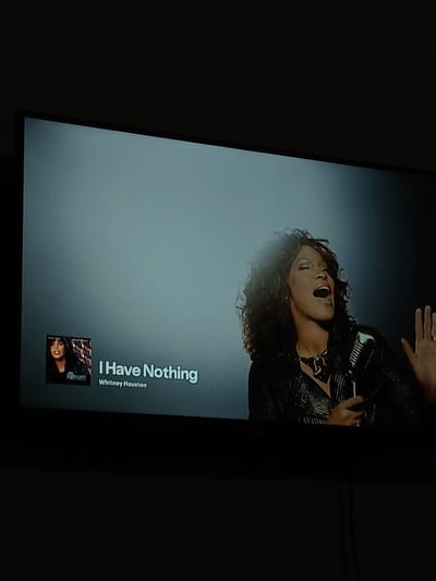 A television screen displays the music video for Whitney Houston's song "I Have Nothing". Whitney Houston is depicted singing into a microphone, with her eyes closed and her mouth open. She is wearing a shimmering black jacket and a statement necklace. To the left of the screen, a small album art image shows a picture of Whitney Houston. The text "I Have Nothing" and "Whitney Houston" are displayed below the album art. The background of the screen is a gradient of light gray to dark gray, with a spotlight effect illuminating Whitney Houston. The television is positioned in a dark room. The image does not provide any information about the location in the city of El Espinal, Venezuela.