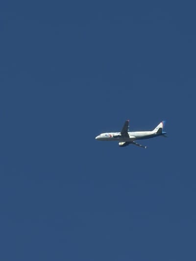 An airplane is visible in the sky, flying at a distance against a clear blue backdrop. The aircraft is white with a blue stripe along its fuselage and red accents on its tail fins. It appears to be in flight, with its landing gear extended. The scene is devoid of any other discernible objects or structures, suggesting an open sky environment. The specific location in Malakhovka, Russia, is not directly identifiable from the image itself.