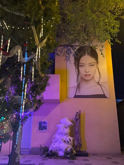 The image shows a street scene, potentially in Linghe, China, though the city's presence is inferred. A large advertisement featuring an Asian woman is mounted on a light-colored building. An umbrella leans against the wall nearby, positioned over a pile of white cushions and foliage. Below this arrangement, a framed picture and a white garage door are visible. In the foreground, a decorated tree with lights, ribbons, and ornaments dominates the lower portion of the frame. Another, smaller tree partially obscures the left side of the building. The overall lighting is dim, with yellow and purple hues illuminating the scene.