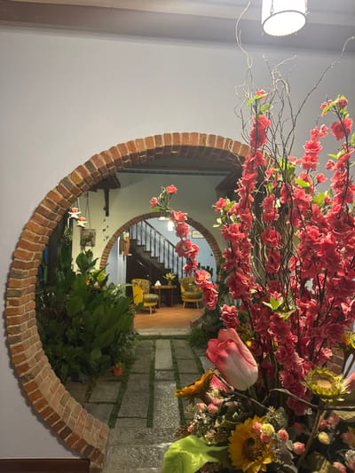 The scene depicts an interior view in George Town, Malaysia. A circular archway, constructed from reddish-brown bricks, frames a path leading into a courtyard. The path consists of rectangular stone slabs set within a darker ground, flanked by dense green foliage. Beyond the courtyard and its plants, a secondary archway opens to an interior room. This room contains a dark wooden staircase with white risers ascending towards the left, and on the right, two yellow armchairs with floral upholstery are positioned around a small, circular wooden table. The floor of this inner room is a reddish-brown tile. In the immediate right foreground, a large, elaborate floral arrangement is prominently displayed. It features numerous bright pink artificial blossoms on dark branches, a pink and white tulip, and several yellow sunflowers, resting in a dark base with a light green fabric drape. An illuminated cylindrical light fixture is visible at the top right of the frame. No individuals are present in the scene.