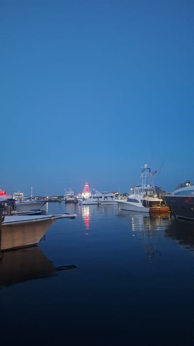This is a vertical, low-angle shot of a harbor at dusk in Nantucket, Massachusetts, USA. The sky is a deep, clear blue. Several boats of various sizes are docked or anchored in the water, their reflections shimmering on the calm surface. In the background, a prominent red and white structure, likely a lighthouse or a similar landmark, is illuminated with red lights. The lights of the boats and the background structure are also reflected in the water. The foreground features the hull of a boat, partially submerged, creating a dark, abstract shape in the water. The overall atmosphere is tranquil and serene, with the fading light casting a soft glow over the scene.