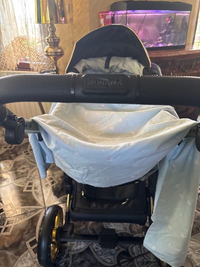The image depicts a close-up view of a baby stroller, specifically the handlebar and the fabric liner within the stroller. The handlebar is black with a textured grip and features a "JUNAMA" logo. A light blue fabric liner with a pattern of pale pink and white circles and stars is draped over the stroller's frame. The background shows a living room environment with a decorative lamp on the left and a fish tank on the right, suggesting an indoor setting. The floor is tiled with a patterned design. There are no people visible in the image. The scene is static and offers no information about any specific location in the city of Slanchevo, Bulgaria.