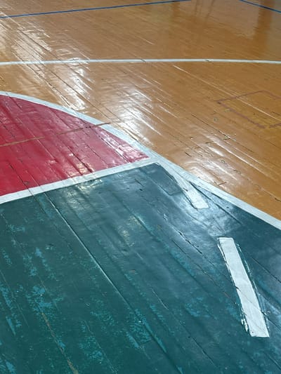 The image displays a close-up, high-angle view of a heavily worn wooden sports court floor, identified as being located in Tayshet, Russia. The surface exhibits extensive degradation, with widespread cracking, chipping, and peeling of paint and varnish across the floorboards. The court features a reddish-brown main area, delineated by a thin blue line in the upper background and faint, faded rectangular outlines suggesting prior markings. This reddish-brown section transitions into distinct red and dark green (teal-colored) painted zones, separated by worn white boundary lines. Both the red and dark green sections show significant paint loss and chipping, revealing the underlying wood grain. A prominent, elongated white marking is present on the dark green section, overlaying a broader, irregularly shaped white area. The floor's glossy, reflective patches indicate an indoor setting with overhead illumination. No individuals, objects other than the floor itself, or specific actions are depicted.