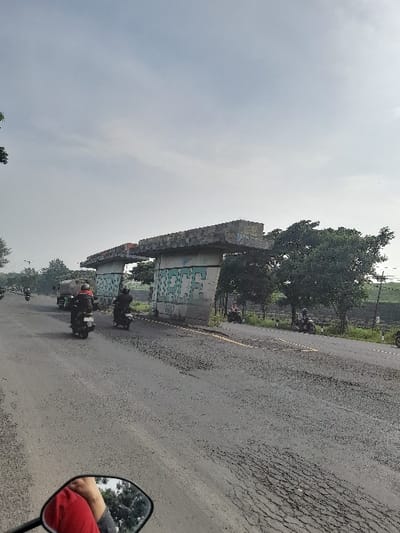 This image captures a daytime scene from the perspective of a person on a motorcycle, looking along a road in what appears to be Kecamatan Gempol, Indonesia. The sky above is overcast and hazy, giving a soft, diffused light to the scene.
The main subjects are a road, a large concrete structure, and several motorcycles. The asphalt road, which stretches diagonally across the frame, shows significant signs of wear with extensive cracking and unevenness. On the left side of the road, a substantial, unfinished or dilapidated concrete structure, possibly part of an overpass or bridge, runs parallel to the road. Its rough surfaces are covered with green graffiti, featuring stylized block letters that are difficult to decipher precisely but might spell out "K.D." or similar.
Several motorcycles with riders are visible on the road, suggesting regular traffic or commuting activity. In the immediate foreground on the right, a rearview mirror reflects the arm and shoulder of the person taking the photo, who is wearing a red shirt, confirming their position on a motorcycle. Beyond the concrete structure and along the road, green trees and dense foliage are present, blending the infrastructure with natural elements. The overall setting portrays an everyday, functional landscape in a semi-urban or developing area.