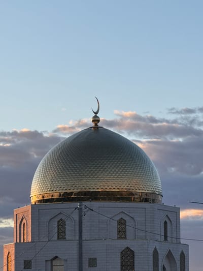This media file shows a close-up, low-angle shot of the dome and minaret of a mosque, with a cloudy sky in the background. The dome is large, covered in a patterned metallic material that reflects the light, giving it a golden hue. A crescent moon sits atop the dome, a common Islamic symbol. Adjacent to the dome is the side of the mosque building, constructed from light-colored stone blocks. The building features decorative arched windows with intricate geometric patterns. Power lines are visible, crossing the frame in front of the building.

The lighting suggests it is either sunrise or sunset, with warm, golden light illuminating the dome and a few streaks of pinkish light in the clouds. The sky is a mix of clear blue and various shades of gray and purple clouds, indicating dynamic weather conditions.

The specific subject matter, the dome with a crescent, and the architectural style are indicative of Islamic religious architecture. Given the location context of Bolgar, Russia, this is likely a prominent mosque in that area. No people are visible in the frame. The overall mood is one of serene grandeur, highlighting the beauty of the religious structure against the dramatic sky. No text is visible in the image.