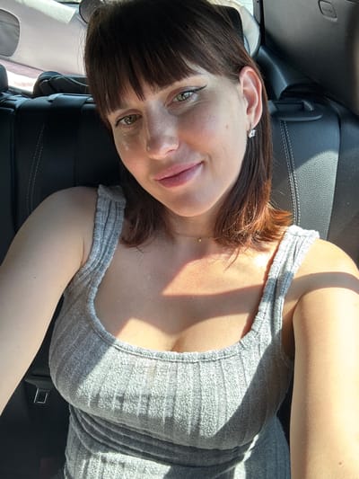 A young woman with brown hair and bangs is reclining in a car seat. She is wearing a grey, ribbed tank top. The interior of the vehicle includes a black leather seat. The location is not discernible as Pomezia, Italy.