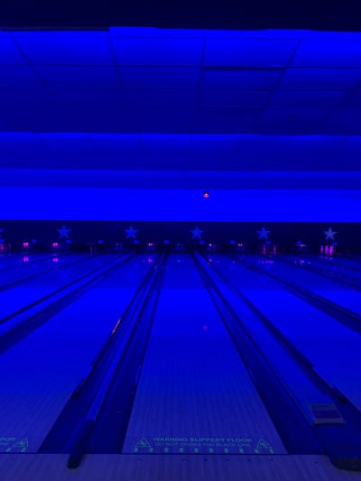 The image shows an empty bowling alley in Bradford, UK, located at latitude 53.791, longitude -1.747. The bowling lanes are illuminated with bright blue lights. The ceiling has a grid pattern and is also lit with blue lights. There are five stars on the back wall, which are also illuminated with blue light. The image shows a close-up of the bowling lanes with a "WARNING SLIPPERY FLOOR DO NOT CROSS THE BLACK LINE" sign at the bottom of the frame. There are no people present in the image.