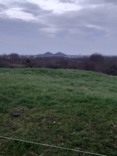 A landscape features a grassy field on the right side, extending into the distance. Beyond the field, a treeline with some buildings and faint lights can be seen. Further in the background, two distinct pyramidal hills or peaks are visible. The sky is overcast and cloudy. A thin, white line diagonally crosses the foreground of the field.