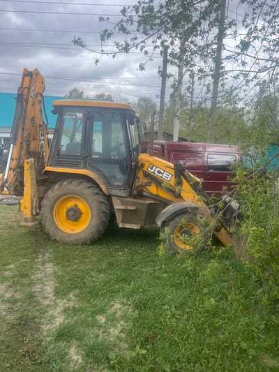 A yellow JCB backhoe loader is parked on a grassy area near a red truck in Поедуги, Russia. Overhead power lines and a cloudy sky are visible above the scene. The area has trees and vegetation.