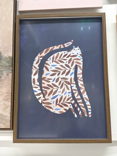 The image shows a framed piece of art featuring a stylized cat against a dark blue background. The cat is depicted in a light color and is covered in a pattern of brown leaves and blue dots. The cat's tail is curved upwards, and its body appears elongated and somewhat abstract. The artwork is housed in a simple wooden frame. To the left of this artwork, a portion of another framed artwork is visible, which appears to be a landscape with muted colors. Above the cat artwork, a pink object, possibly another piece of art or decorative item, is partially visible. The setting appears to be an indoor space, potentially a gallery or a home, with white walls and lighting that causes some reflections on the surface of the artwork. The specific location in the city of Son en Breugel, Netherlands, is not discernible from the visual information provided.