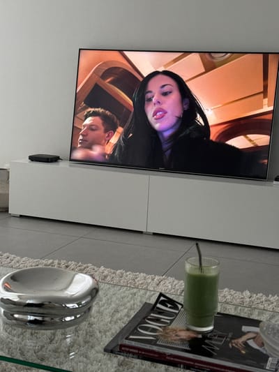 The scene depicts an indoor living space, located in Tielrode, Belgium. A large, flat-screen Sony television is centrally positioned on a white, modern two-door media console. To the left of the television, a small black electronic device rests on the console.
The television screen displays a broadcast featuring two individuals in an interior setting with warm, orange-toned architectural elements. A female individual with long dark hair, fair skin, and full lips is prominent on the right side of the screen, looking slightly towards the viewer with her mouth slightly open. She wears a dark garment, possibly faux fur, and a small hoop earring. Behind her and to the left, a male individual with short dark hair and fair skin is partially visible, looking away from the camera. His bare shoulder or light-colored top is visible.
In the foreground, a clear glass coffee table sits on a light-colored shag rug. On the table, a reflective silver-toned, organically shaped decorative bowl or dish is present. Adjacent to it, a "VOGUE" magazine with a woman featuring blonde hair on its cover lies open. A ribbed glass containing a green liquid and a dark straw is placed on top of the magazine. The floor beneath the rug and console is composed of large, light grey square tiles. The wall behind the media console is plain white.