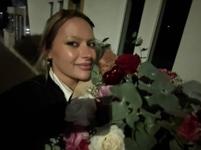A person is shown holding a bouquet of flowers. The flowers include a mix of white, pink, and red roses, along with eucalyptus leaves. The individual is wearing a white collared shirt under a dark blazer. They have blonde hair and are smiling. The background appears to be an interior space with windows, possibly a building. The image is a bit dark and taken from an unusual angle.