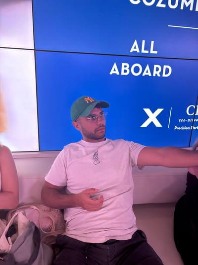 The image displays an indoor scene, likely within a ferry terminal in San Miguel de Cozumel, Mexico. A male individual, approximately 20-30 years old, is seated centrally on a light-colored bench. He wears a green baseball cap with an orange "NY" logo, round wire-frame glasses, a light-beige t-shirt with a black minimalist graphic of a head emitting plants, and dark grey drawstring pants. He possesses facial hair, including a beard and mustache, and is looking towards his left. His right arm is extended horizontally to the right of the frame. To his left, a female individual is partially visible, seated, next to a collection of bags including a mesh-style tote. To his right, another partially visible individual is seated. In the background, a large, multi-panel blue digital display features white text: "COZUMEL" (partially visible), "ALL ABOARD", a large "X", and smaller text indicating "Eco-Drive" and "Precision Platform," suggestive of a modern transportation service. The environment is illuminated by artificial light.