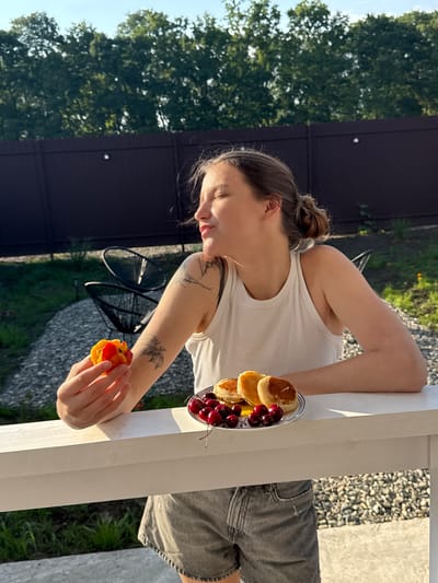 A young woman with her hair in a bun leans against a white railing outdoors. She wears a white tank top and grey denim shorts. A small plate with mini pancakes and cherries sits on the railing in front of her. She holds a piece of fruit, possibly a peach or nectarine, in her right hand. She has tattoos on her left arm. The background shows trees, a dark fence, and gravel. The sun is shining, casting shadows, suggesting it is daytime, possibly late afternoon. The setting appears to be a backyard or patio area. The woman's facial expression is relaxed and she seems to be enjoying her snack. No visible text.