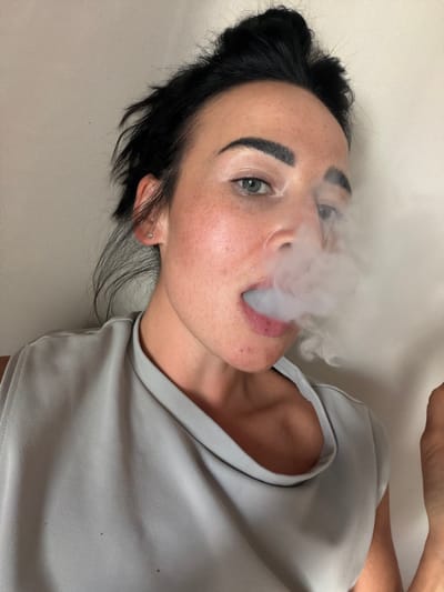 A close-up, vertically oriented photograph shows a woman with dark hair and light eyes exhaling a plume of white smoke. Her hair is pulled back, revealing her ear with a small stud earring. She has prominent eyebrows and a smattering of freckles across her nose and cheeks. Her lips are slightly parted, and the smoke billows from her mouth. She wears a light grey t-shirt. The background is a plain, off-white surface, suggesting an indoor setting. The lighting is soft, and there are no discernible time-of-day or weather cues. The image appears to have been taken in Koltushi, Russia.