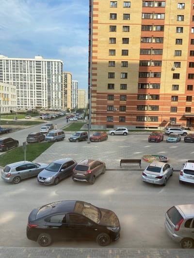 Here's a detailed description of the image:
**Setting:** The image appears to be taken from a high vantage point, likely from an upper floor of a building in Дядьково, Russia. The buildings in the foreground and background suggest a dense urban residential area. The sky is partially visible with a blue hue indicating a clear day.
**Buildings:** There's a large, multi-story apartment building with a brick facade consisting of alternating bands of yellow and red brick. Other residential high-rise buildings are visible in the distance, colored primarily in white.
**Transportation:** A street runs between the buildings, packed with parked cars. The street is paved in grey tarmac.
**Vehicles:** Numerous cars are parked along the sides of the road, facing different directions. A silver-gray sedan stands out in the foreground, partially off-frame. Car colors include silver, red, brown, gray, and white.
**Other Objects:** A small children's playground with a sandpit is visible next to one of the buildings. A metal bench is seen next to the playground. Patches of grass are noticeable between parked cars along the side of the road.
**People:** No people are clearly visible.