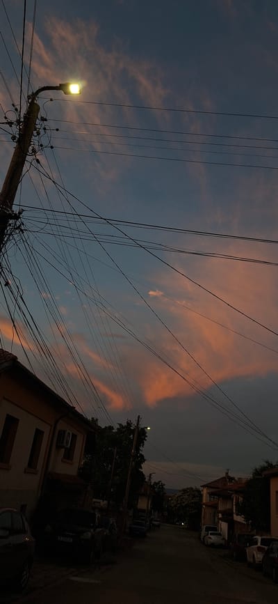 The image depicts a street scene at dusk in Lyaskovets, Bulgaria. The sky is a dusky blue with streaks of pink and orange clouds, illuminated by a bright street lamp. A wooden utility pole, heavily laden with tangled wires, stands on the left side of the frame, casting shadows. Adjacent to the pole is the side of a building with a light-colored facade, featuring windows and an air conditioning unit. The street is lined with parked cars, some partially visible in the dim light. Further down the street, more buildings and trees are visible, with a hint of distant hills. The overall atmosphere is somber and quiet, characteristic of the end of the day in a residential area.