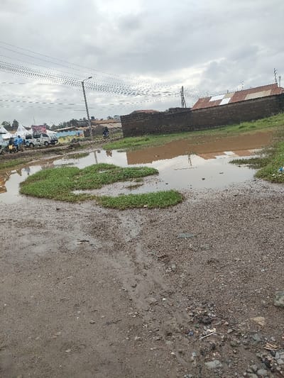 The image depicts an outdoor scene in Juja ward, Kenya, under an overcast sky. The foreground and mid-ground consist of saturated, unpaved ground composed of mud and gravel, featuring several large, irregularly shaped puddles of murky brown water. Patches of green grass grow within and alongside these water bodies. To the right, a low, dark brown brick or stone wall fronts a building with a corrugated metal roof, presenting sections of white and rusted reddish-brown. Multiple antenna-like structures are visible along the roofline. In the left background, a cluster of white tent-like structures and other low-rise buildings are visible. Two white utility vehicles, possibly vans, are parked near these structures. Several individuals are present in the vicinity of the vehicles and tents, their reflections indistinctly visible in the foreground puddles. In the mid-ground, a person on a motorcycle is observed moving away from the viewer on a muddy path. A tall utility pole with multiple overhead power lines stretches across the mid-ground, exhibiting a slight lean. The ground throughout displays evidence of vehicle tracks and scattered small debris.