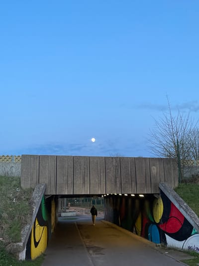 This image depicts a concrete underpass structure in the city of Plaisir, France, observed during twilight hours with a clear, light blue sky overhead and a prominent full moon visible in the upper-middle sky. The underpass, constructed from vertically arranged concrete panels on its upper surface, features internal walls adorned with vibrant, abstract graffiti in red, yellow, green, and blue hues. Overhead lighting fixtures illuminate the paved pathway running through the tunnel.

One individual, wearing a dark jacket and light-colored trousers, is walking away from the viewer along the pathway inside the underpass, moving towards the distant exit. Beyond the underpass, two indistinct figures are visible on a paved area, one standing and one closer to the ground. The surrounding environment includes grassy slopes leading up to the underpass structure, with some visible litter (white specks) on the left slope. Bare deciduous trees and bushes are present on the right side, behind the underpass. A yellow lattice fence is situated on the top left, and another wire fence is visible on the right side.
