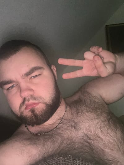 A close-up, low-angle, chest-up selfie of a man with a beard is shown. The man is looking at the camera with his left eye partially closed and his mouth slightly open. He is wearing a silver chain around his neck. His right hand is raised, with his index and middle fingers extended in a peace sign. The background is a dark, textured wall. The man appears to be indoors, possibly in a bedroom. There is no specific information to place the image in Gelsenkirchen, Germany.