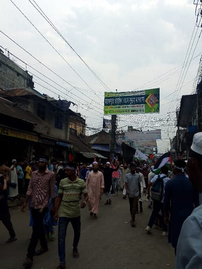 The image shows a street scene in Bhola, Bangladesh (22.686, 90.644). A large group of people are walking down the street, mostly men. Some are wearing traditional clothing, including white shirts and pants, and white skullcaps. Several men are wearing long, loose white robes. There are several flags hanging on buildings and being carried by individuals. The flags appear to be green and white, and one has a red triangle on the left. There is a large banner hanging from a pole in the middle of the street, written in the Bengali language. The banner is green and white with a red triangle. There are many power lines running across the image. The buildings lining the street are mostly low-rise, with some businesses on the street level. There are a few trees on the street, and the sky is cloudy. The people in the image appear to be walking in the same direction, and there are some conversations happening among them. The scene appears to be a public gathering or protest march.