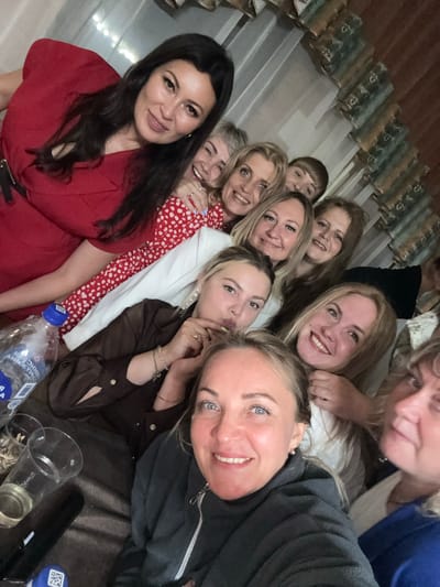 The image shows a group of ten women posing for a selfie in what appears to be an indoor gathering. They are all smiling and looking at the camera. The women are of various ages and ethnicities, and they are dressed in casual clothing. Some of the women are wearing dresses, while others are wearing tops and pants. There are a few drinks and a bottle of water on a table in the foreground. The background is blurred, but it appears to be a room with some decorations on the wall. The setting is consistent with a private celebration or casual get-together, possibly in Zavod Mikhailovskii, Russia, given the context provided.