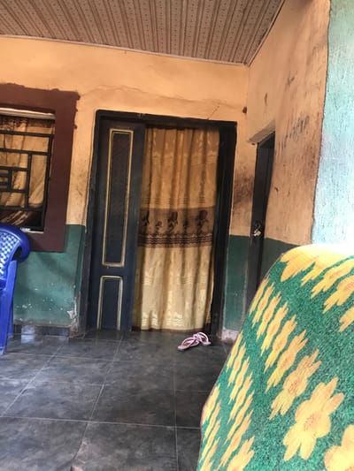 A residential entry presents a dark door with a curtain. A smaller, grated window sits below it. To the right, dark gray tiled flooring with a pair of pink flip-flops is partially covered by a green and yellow floral mat. A blue plastic chair is visible near the bottom. Walls are painted in varying colors including beige, green and blue. The ceiling appears unfinished.