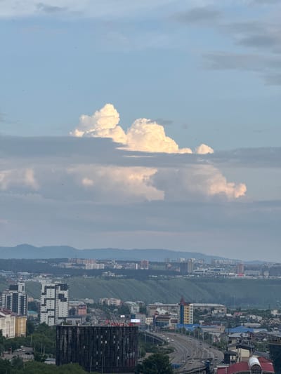 The image depicts an aerial view of Ulan-Ude, Russia, from a high vantage point. The scene is dominated by a vast sky filled with dramatic clouds, some of which are brightly lit and fluffy, while others are dark and overcast. Below the sky, a sprawling cityscape unfolds, characterized by a dense collection of buildings nestled against a backdrop of lush, green hills. A prominent highway with traffic can be seen winding through the city.

The time of day appears to be late afternoon or early evening, as indicated by the lighting conditions and the quality of the sunlight illuminating the clouds. The weather seems mixed, with patches of clear sky interspersed with heavy cloud cover. The city itself displays a mix of architectural styles, with modern high-rises and older, more traditional structures. The presence of a highway and numerous residential and commercial buildings suggests a populated urban environment.

No people are clearly discernible in the image, and the focus is on the expansive landscape and the urban sprawl. The overall mood of the image is somewhat dramatic, due to the interplay of light and shadow on the clouds and the sheer scale of the cityscape against the natural landscape. No visible text is present within the image.