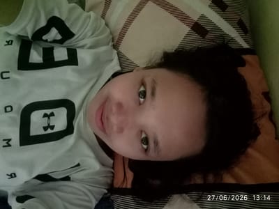 The image shows a young child lying on a bed, wearing a white t-shirt with black lettering and a logo. The child's head is tilted to the side, and their eyes are open. The background consists of a striped pillow and a green wall. The image was taken indoors, likely in a residential setting in Sukaraja, Indonesia, as indicated by the timestamp. There are no other people or significant objects in the frame.