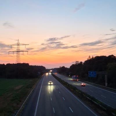 The image depicts a highway scene at dusk, with a gradient sky of pale blue fading into orange and pink hues. A tall electricity pylon stands prominently on the left, silhouetted against the colorful sky, with power lines stretching across the frame. Dense, dark trees line the sides of the highway. Several vehicles are visible on the multi-lane road, their headlights illuminated, suggesting the approaching night. A large blue road sign on the right indicates "A 5 Rastatt-Sud 1000 m". The overall environment is a rural or semi-rural highway setting in Germany, likely near Baden-Baden given the sign. There are no visible people.