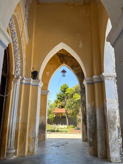 The image depicts an arched passageway, likely part of an old building, with a view of an outdoor scene through the arch. The architecture features a pale yellow, textured wall and decorative elements in white and blue. The arch itself is curved and pointed at the top. Above the arch, there are several horizontal concrete beams with a weathered appearance. Decorative white moldings with floral motifs are visible along the lower edges of the arch and the beams. A small black speaker is mounted on the yellow wall above the arch.

Through the arch, a bright blue sky is visible, partially obscured by lush green trees. In the distance, a building with a reddish-tiled roof can be seen. Hanging from the ceiling within the arch is a decorative, cage-like object, possibly a lamp or a lantern. The overall scene suggests a warm, sunny day, with the light casting shadows and highlighting the textures of the aged structures. The location context provided is Hội Tín, Vietnam, which could imply a historical or cultural site. No people are visible in the image.